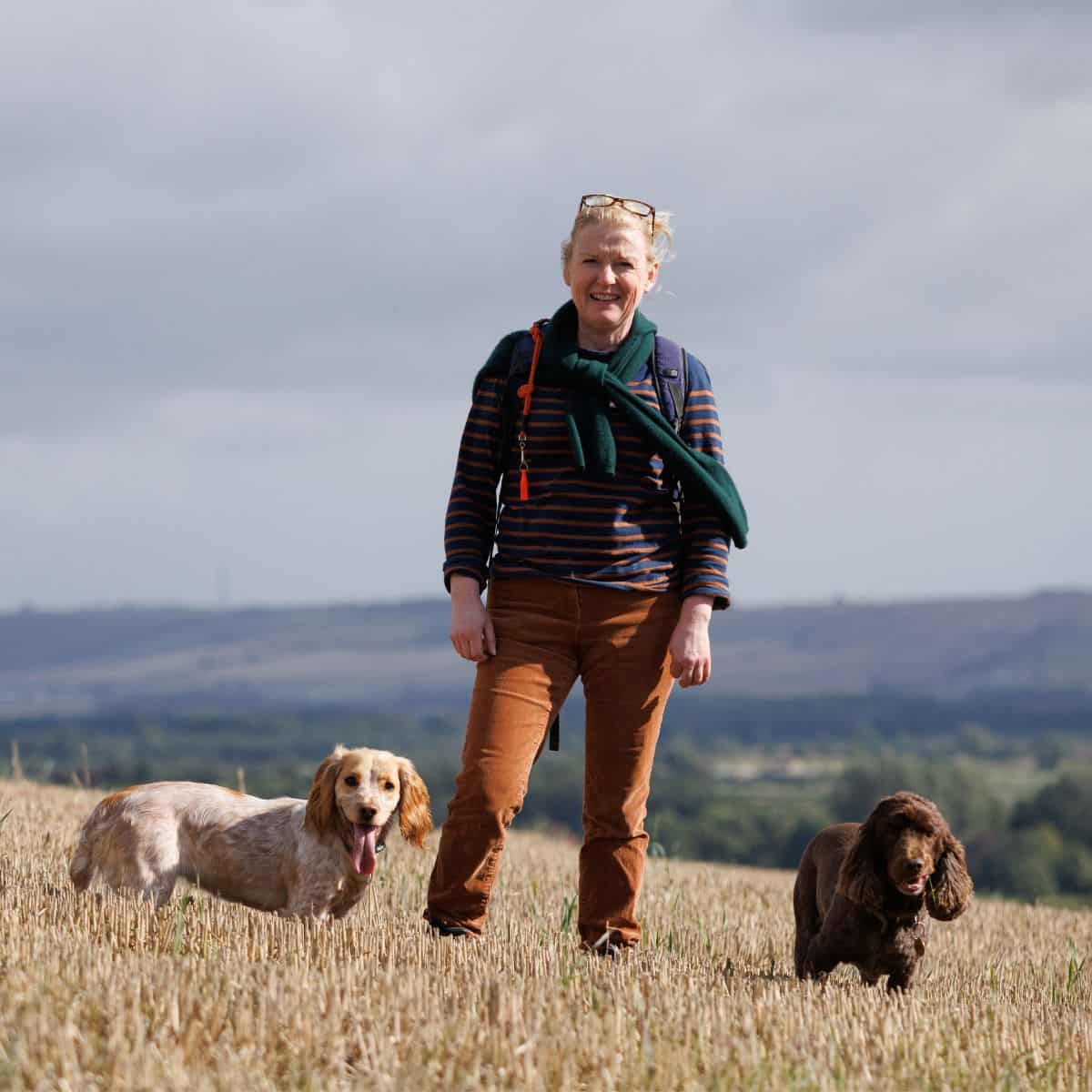 Bright woman hiking outdoors with two dogs, scenic countryside background, active outdoor lifestyle representation.